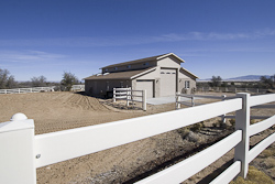 High-end real estate photography exterior view of a modern ranch-style horse barn with stable doors and surrounding wooden fencing in Williamson, AZ. by Tom Hunter Photography